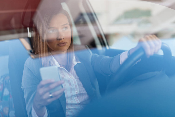 Beautiful businesswoman driving to work and using cell phone behind the wheel. The view is through the glass.  - Fotoğraf, Görsel