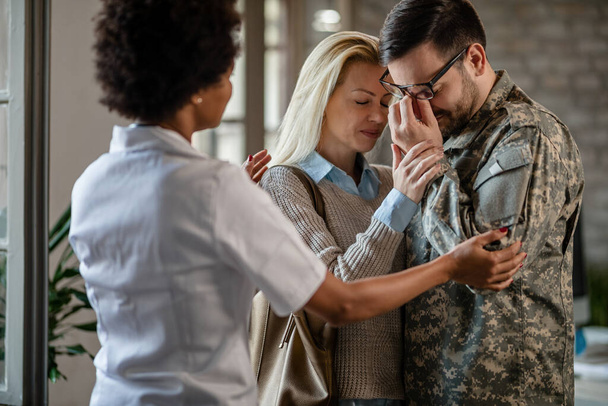 Sad military man and his wife crying after receiving bad news while African American doctor is consoling them at medical clinic.  - Foto, imagen