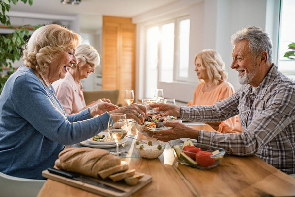 Счастливый старший мужчина передает салат своей жене, наслаждаясь обедом со своими друзьями дома.  - Фото, изображение