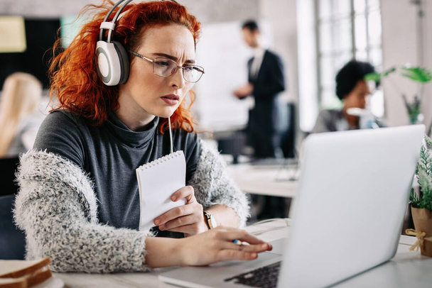 Young casual businesswoman reading problematic e-mail on laptop while listening music on headphones at work. There are people in the background.  - Foto, afbeelding