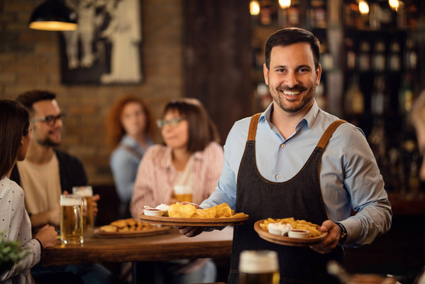 Счастливый официант держит тарелки с едой и смотрит в камеру, обслуживая гостей в ресторане.  - Фото, изображение