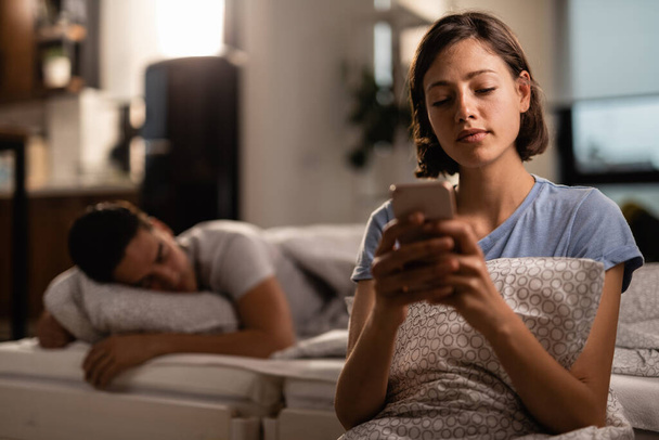 Νεαρή γυναίκα στέλνει μηνύματα στο κινητό ενώ ο φίλος της κοιμάται στο κρεβάτι πίσω της..  - Φωτογραφία, εικόνα