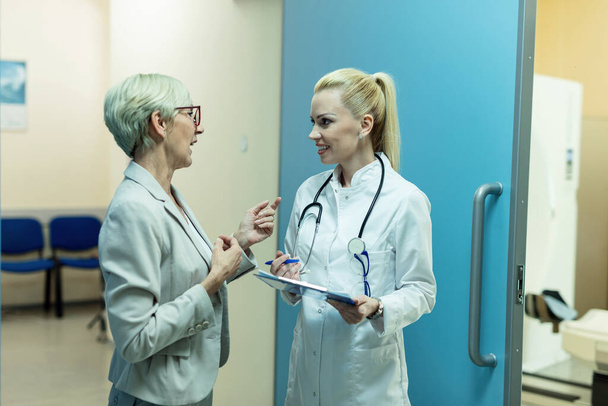 Ευτυχισμένη γυναίκα γιατρός επικοινωνεί με τον ανώτερο ασθενή, ενώ στέκεται σε ένα διάδρομο στην κλινική.  - Φωτογραφία, εικόνα