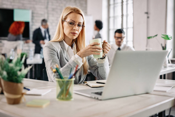 Серйозна бізнес-леді п'є чай і розмірковує під час роботи в офісі. На задньому плані є люди
.  - Фото, зображення