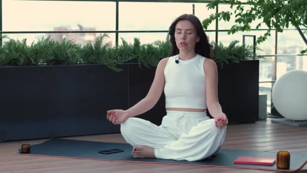 Beautiful woman in white clothes sits cross-legged on karemat on terrace with greenery, practices meditation with closed eyes, feels inner harmony and balance, improves self-awareness, stress-free - Filmagem, Vídeo