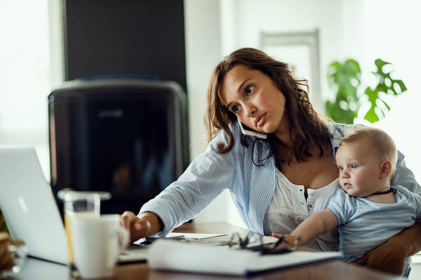 Young mother holding her baby while working on a computer and communicating over mobile phone at home.  - Photo, image