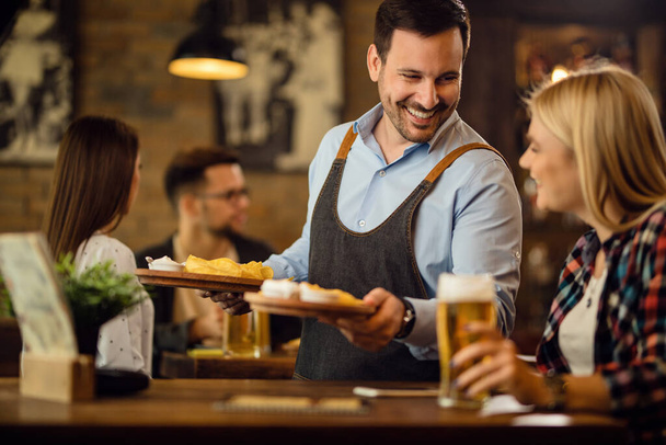 Happy waiter serving tortilla chips to female guest while she drinking beer in a tavern.  - Photo, Image