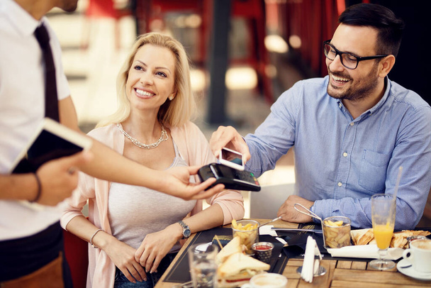 Ευτυχισμένος άνθρωπος κάνει εύκολη πληρωμή με το κινητό τηλέφωνο, ενώ έχοντας ένα γεύμα με την κοπέλα του σε ένα μπαρ.  - Φωτογραφία, εικόνα