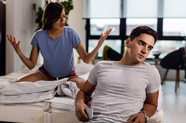 Young man sitting by the bed and ignoring his girlfriend who is arguing with him.  - Foto, Bild
