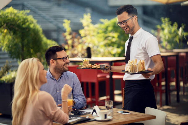 Молодой улыбающийся официант обслуживает пару и приносит им еду во время обеда в ресторане.  - Фото, изображение