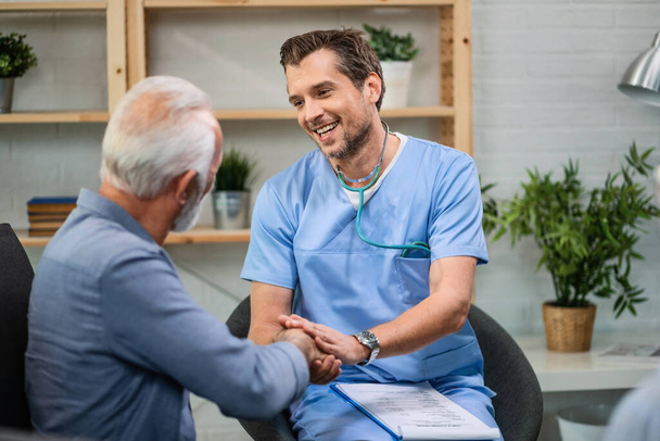 Happy general practitioner talking to senior man while shaking hands with him during a home visit.  - Φωτογραφία, εικόνα