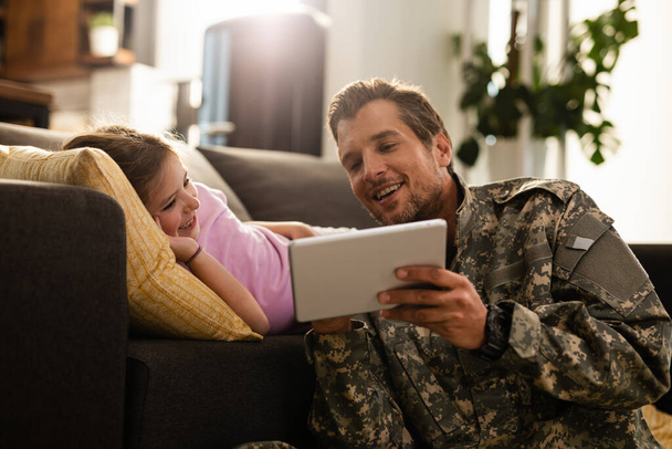 Щаслива дівчинка лежить на дивані і дивиться щось на тачпад зі своїм військовим батьком
.  - Фото, зображення