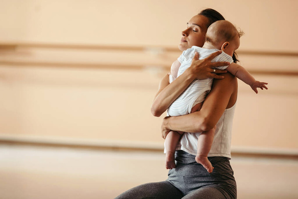 Νεαρή αθλητική μητέρα αγκαλιάζει το μωρό της με τα μάτια κλειστά σε ένα γυμναστήριο. Αντιγραφή χώρου.  - Φωτογραφία, εικόνα