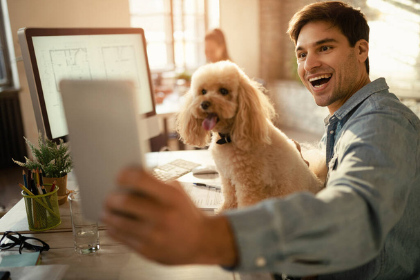 Щасливий бізнесмен використовує цифровий планшет і приймає селфі зі своїм пуделем під час роботи в офісі
.  - Фото, зображення