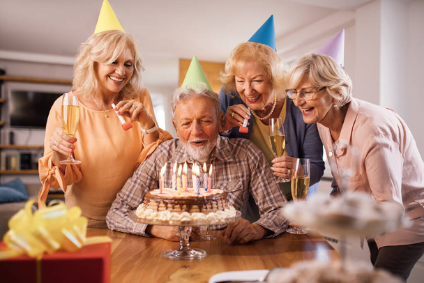 Щасливий старший чоловік дме свічки на день народження під час святкування з друзями вдома
. - Фото, зображення
