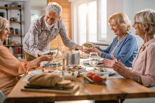 Cheerful senior man serving food to his female friends at dining table at home.  - Foto, Bild