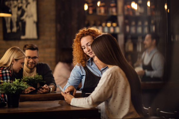 Молода офіціантка допомагає жінці з вибором замовлення з меню в барі
. - Фото, зображення