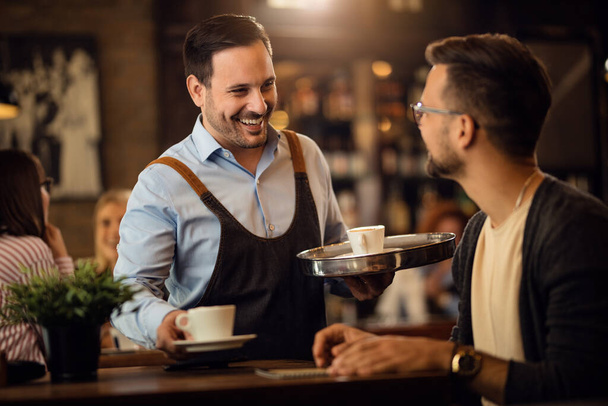 Счастливый официант, подающий кофе и общающийся с гостями-мужчинами в баре.  - Фото, изображение