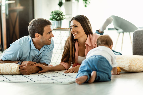 Χαμογελώντας γονείς χαλαρώνοντας στο πάτωμα και μιλώντας μεταξύ τους, ενώ περνούν το χρόνο τους με το γιο τους στο σπίτι.  - Φωτογραφία, εικόνα