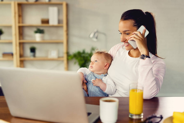 Young happy woman communicating over mobile phone while relaxing with her baby at home.  - Foto, Bild