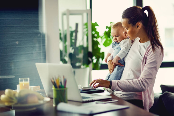 Young working mother using computer while holding her baby son at home.  - Foto, Bild