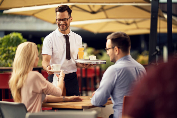 Молодой счастливый официант приносит кофе и напитки гостям в кафе на открытом воздухе.  - Фото, изображение