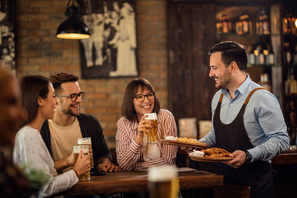 Группа счастливых молодых людей пьет пиво, пока официант приносит еду за их столом в таверне.  - Фото, изображение