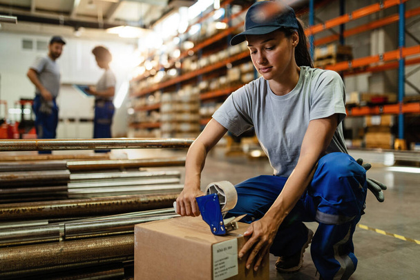 Young warehouse worker preparing cardboard boxes for shipment while using tape dispenser in industrial warehouse.  - Foto, Bild