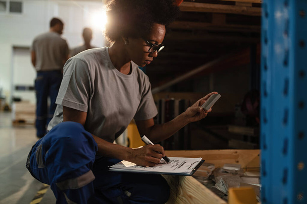 African American female worker checking products before the shipment and taking notes in a warehouse.  - Фото, изображение