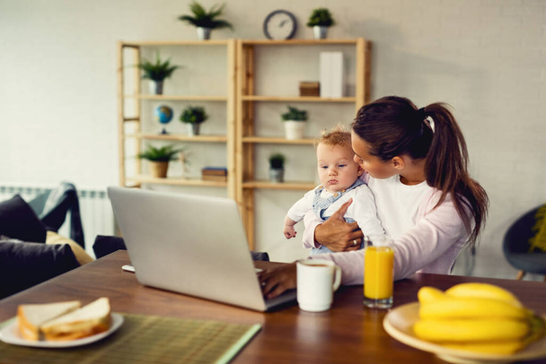 Young mother holding her baby boy while working on laptop at home.  - 写真・画像