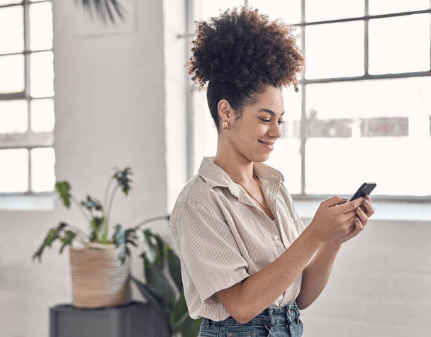 Young cheerful mixed race businesswoman typing a message on a phone at work. One creative hispanic female businessperson with a curly afro using social media on her cellphone in an office. - Foto, immagini