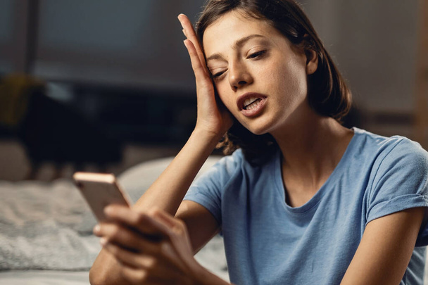 Young displeased woman using smart phone and surfing the internet in the evening in bedroom.  - Foto, afbeelding