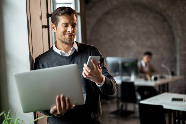Ευτυχισμένος επιχειρηματίας που στέκεται δίπλα στο παράθυρο του γραφείου και διαβάζει γραπτό μήνυμα στο κινητό ενώ κρατάει το laptop.  - Φωτογραφία, εικόνα