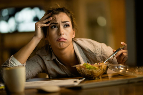 Young distraught woman using computer while eating salad at night at home.  - Foto, afbeelding