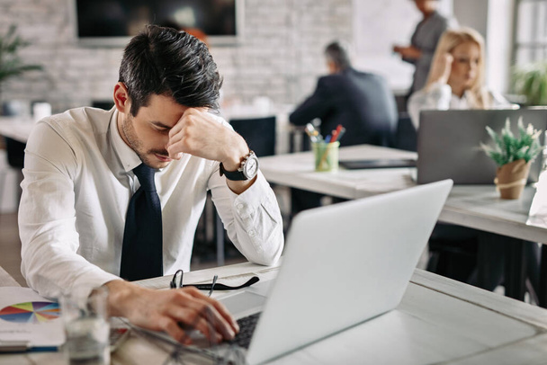 Tired businessman holding his head in pain while working on laptop and feeling exhausted from work. There are people in the background.  - Photo, Image