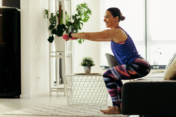 Щаслива спортсменка робить присідання під час тренувань з гантелями у вітальні
.  - Фото, зображення