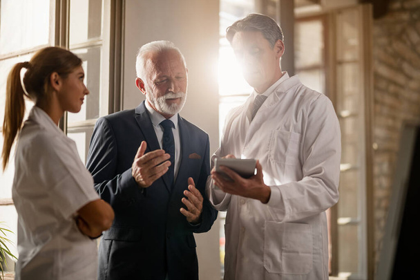 Mid adult doctor showing reports on digital tablet to senior businessman at medical clinic.  - 写真・画像