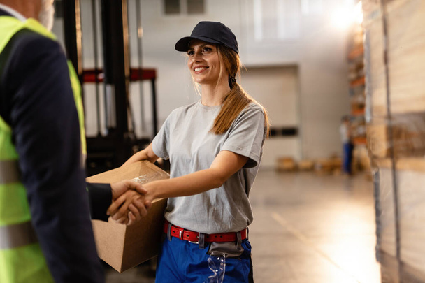 Счастливая женщина-работница приветствует менеджера компании и пожимает ему руку на промышленном складе.  - Фото, изображение