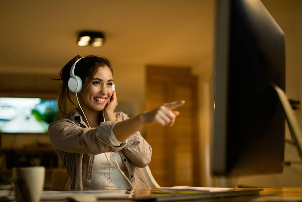 Ευτυχισμένη γυναίκα με ακουστικά που δείχνουν στην οθόνη του υπολογιστή, ενώ σερφάρετε το βράδυ στο σπίτι.  - Φωτογραφία, εικόνα