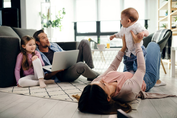 Playful mother holding her baby son while lying on the floor in the living room. Father and daughter are using laptop.  - Foto, Imagen