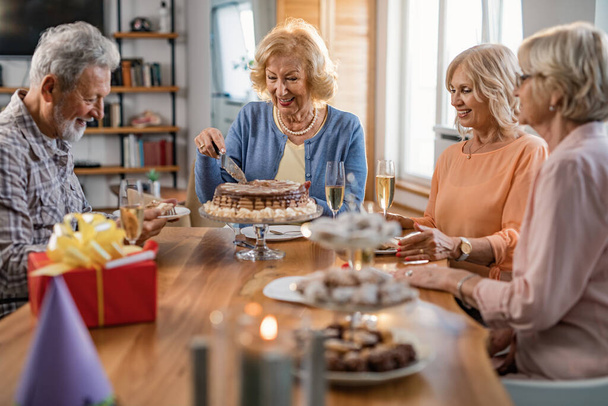 Щасливі літні люди святкують день народження жінки, коли вона ріже торт вдома
.  - Фото, зображення