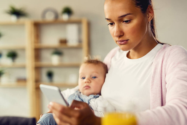 Ψυχαναγκαστική μητέρα χρησιμοποιώντας το κινητό τηλέφωνο και πληκτρολογώντας μήνυμα κειμένου, ενώ είναι με το μωρό της στο σπίτι.  - Φωτογραφία, εικόνα