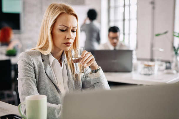 Worried businesswoman working on a computer while drinking water from a glass in the office.  - Foto, Bild