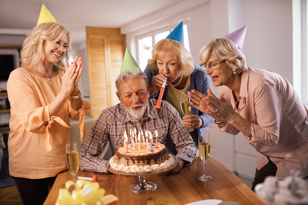 Group of happy senior friends celebrating Birthday at home. Man is blowing candles on a cake.  - Foto, afbeelding