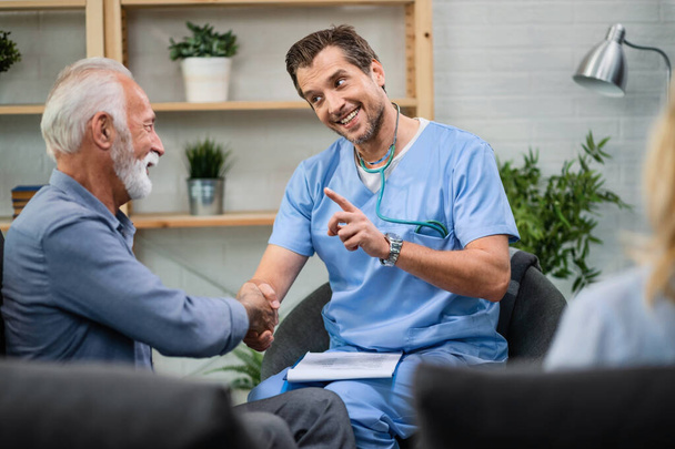 Happy doctor communicating with senior man while shaking hands with him during a home visit.  - Valokuva, kuva