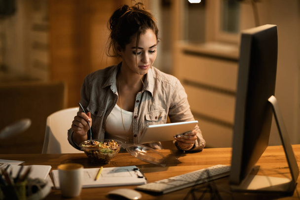 Smiling woman surfing the net on digital table while eating salad in the evening at home.  - Foto, imagen