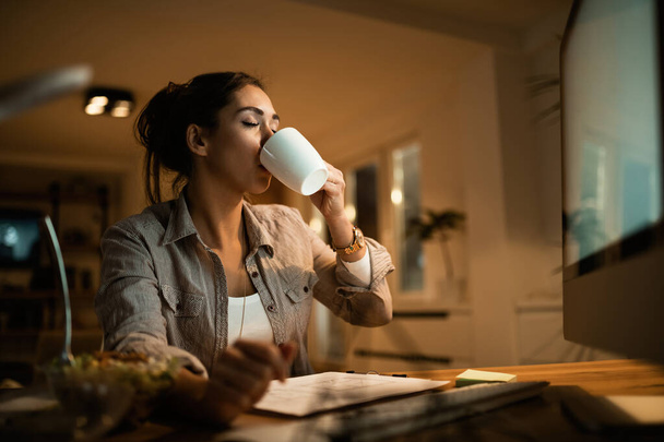 Νεαρή γυναίκα πίνει καφέ με τα μάτια κλειστά ενώ διαβάζει τη νύχτα στο σπίτι.  - Φωτογραφία, εικόνα