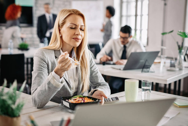 Businesswoman eating healthy and enjoying in taste of vegetable salad with eyes closed during her lunch break.  - Fotó, kép