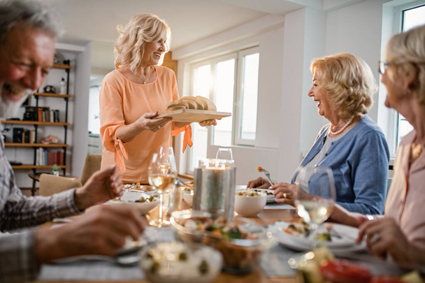 Щаслива старша жінка, що подає хліб, розмовляючи зі своїми друзями за обіднім столом вдома
.  - Фото, зображення