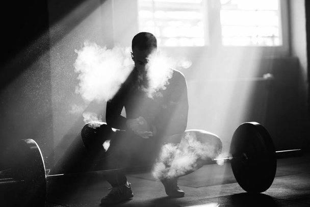 Ασπρόμαυρη φωτογραφία του μυώδους άντρα χρησιμοποιώντας αθλητική κιμωλία om τα χέρια του πριν από την άσκηση με ένα κουδούνι σε ένα γυμναστήριο.  - Φωτογραφία, εικόνα